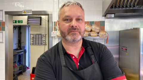 Marcin Wysocki, in his 40s, with grey hair and light stubble, stands in the kitchen of a takeaway wearing a black polo-neck t-shirt and black apron and has his arms folded.