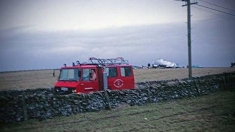 Old faded photo of red rescue vehicle on a road behind a wall in between fields, with the wreckage of a passenger plane in the background.