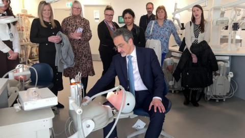 Dr Barrak wears a dark blue suit and tie and sits on a stool astride a dummy head with equipment that will be used to train the students. A group of people behind him watch.