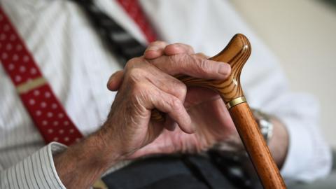 A close-up of an elderly man's hands. He is holding a wooden walking stick and wearing a white pin-striped shirt with red braces.