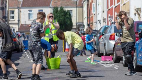 A group of children have a water fight on a suburban street on a sunny day. A woman in a yellow high-vis jacket is supervising. Cars line the street.