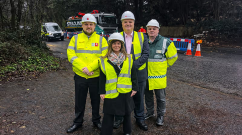 The image shows four people posing for a photograph: they are all wearing hi-vis jackets and there is a road behind them, presumably where the construction is taking place.