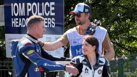 Channing Tatum on the left has blue and black leathers on, he's reaching for a hug to Eve Hewson on the right with black and white leathers on. She has dark hair tied up and is smiling at him. Director Reid Carolin is behind them with his hand on Channing's shoulder, wearing a TT bib over a t-shirt, with headphones over his shoulder, and wearing sunglasses and a TT cap.