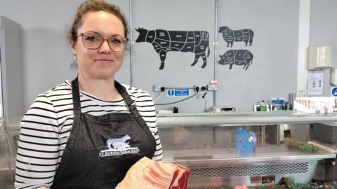 A woman with brown hair tied back is standing in front of a meat counter in a farm shop. She is holding a piece of beef. She is wearing a black apron with the words St Buryan Farm Shop on and a black and white striped Breton top. Behind her on the wall are butchery guides for a cow, sheep and pig.