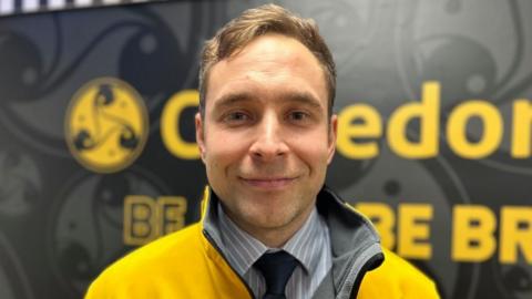 PE teacher Elliot Nightingale, who has short brown hair and is smiling. He is wearing a yellow jacket and is standing in front of a blurry yellow and black sign.