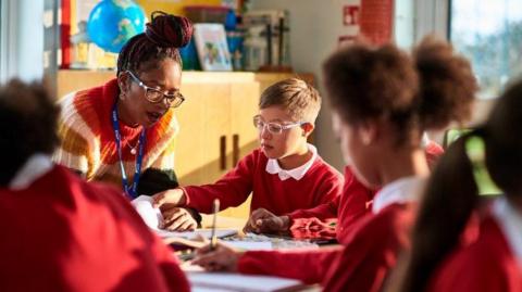 A teacher in a striped multi-coloured jumper looks at a student's book next to a student wearing a red school jumper. They are surrounded by other pupils in the same red uniform.