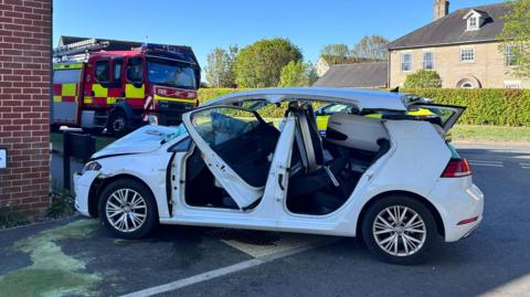 A badly damaged white Volkswagen car parked in front of the corner of a house in which it had crashed into. Its roof is resting on the top of the car having been cut away from the its body, its passenger side door is smashed into the vehicle, and a fire engine is parked behind it.