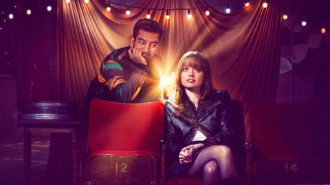 Nabhaan Rizwan and Aimee Lou Wood face towards the camera, a film projector its between them, a bright light illuminating the image.