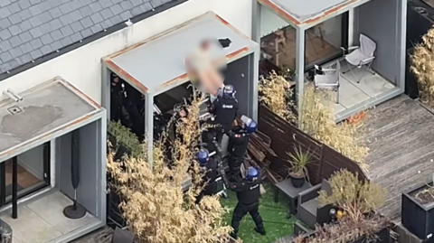 Drone footage shows officers pulling a blurred man from a small roof.