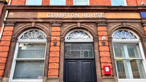 A red brick building with three arch windows and a black door in the centre. A sign for "Compton House" is above the door. 