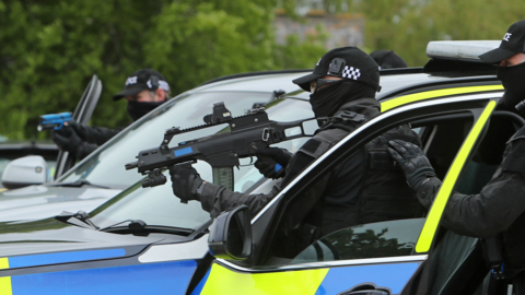 Two masked police officers stood beside a white police car, holding guns 