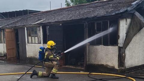 A firefighter in full gear shown hosing down the inside of a low, burned-out structure which appears to be a workshop. 