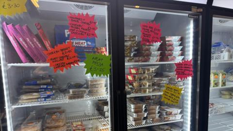 A black and white fridge with food inside. The fridge has multi-coloured stickers with names of foods and prices on them, such as "meat pizza - £3.15".