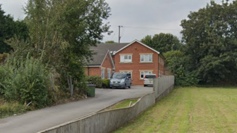 A house at the end of a long driveway. There are trees on one side and a green on the other.