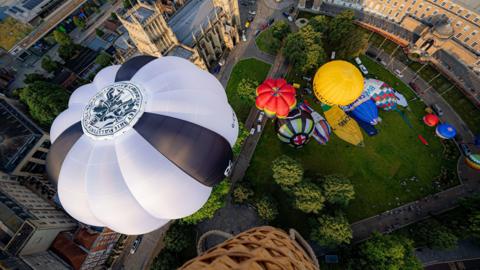 A view from the sky of hot air balloons, white, yellow and red, flying from near Bristol Cathedral above a College Green.