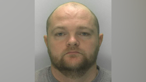 A mug shot of Anthony Williams. He is a balding man with a trimmed beard, wearing a grey jumper