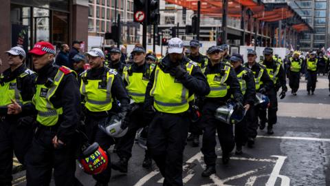 Rows of police in riot gear walking down a street in central London. They are all wearing caps and fluorescent yellow tabards and are carrying riot helmets. 