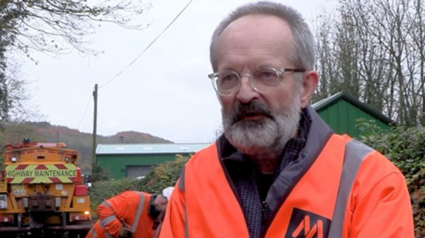 A man with grey hair, a beard and an orange jacket in front of bushes, a man with an orange jacket and a yellow lorry