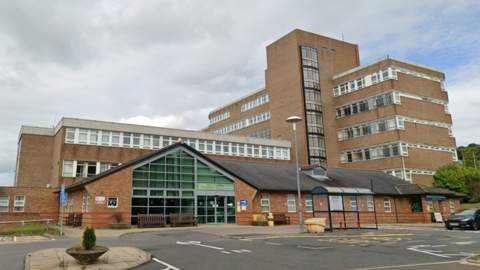 A large brick built hospital with a shelter in front. In front is a large single-storey building with a triangular glass-fronted entrance. Behind it is a three-storey building with long, uninterrupted windows along each floor. To the right is a seven-storey building in the same style, with a large stairwell structure just off-centre. The buildings are arranged in a T-shape with the tallest building at right angles to the other two.