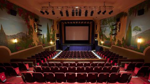 The interior of a theatre facing towards the stage, dimly lit by lights on the wall on either side and with dozens of seats in front of it.