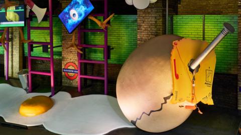 A surrealist art exhibit on a disused Tube platform featuring two giant cardboard eggs, one cracked open with a yellow yolk and the other with a nail sticking out of it. There are oversized ladders, megaphones and large chicken's legs as part of the exhibit. 