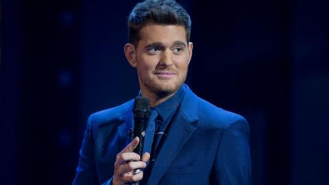 Michael Bublé stands on a dark stage in a royal blue suit, shirt and tie. He is holding the microphone in his right hand, looking to the left nd smiling slightly. He has thick brown hair and stubble.
