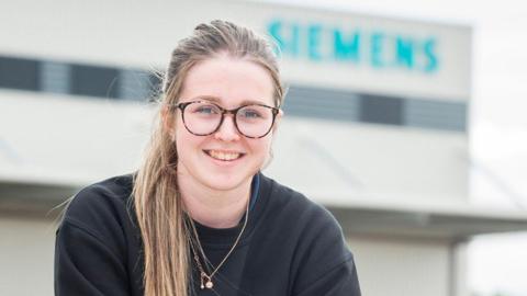 Liv has long blonde hair tied back, has wide rimmed, circular-framed glasses. She is smiling for the camera, with a Siemens sign on the side of a building in the background.