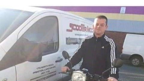 Lee Warburton, with short brown hair, wears a black tracksuit top as he sits on a motorbike parked up near a white van near a warehouse.