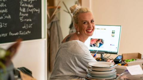 Lucy Sidwick is standing by a till in a cafe with a stack of plates and a computer next to her. She is smiling at the camera.