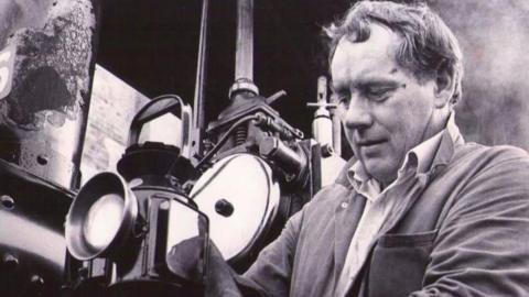 The picture shows a black and white image of William Scurlock in overalls.  He is looking at the machinery he is working on.