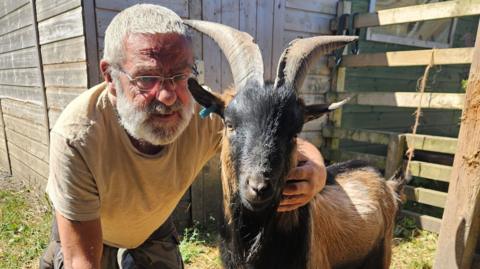 Wayne Eckersley, sitting down, with his arm around a goat looking straight at the camera