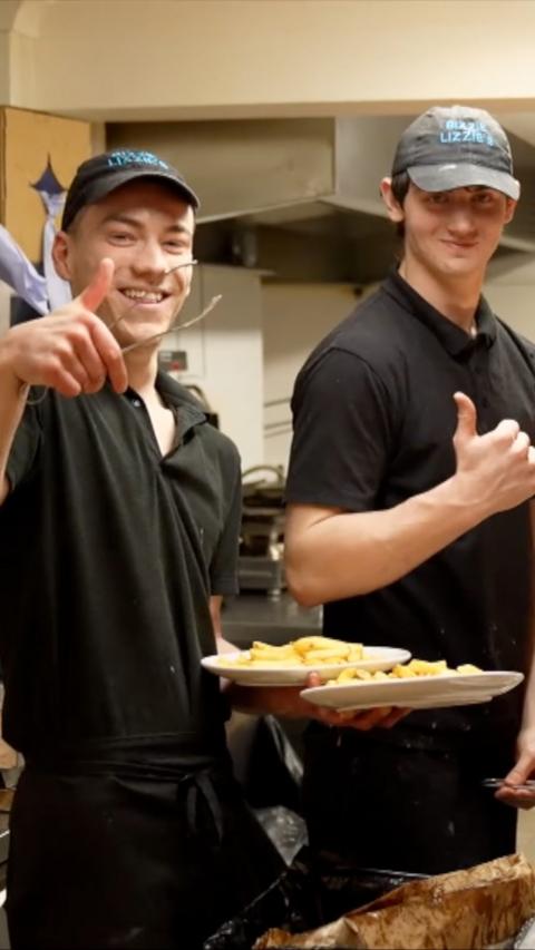 Two chip shop employees in black uniform and caps put thumbs up and smile.