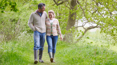 A man with dark hair and a woman with blonde hair both wearing beige jumpers, jeans and wellington boots walking through a grassy area with trees behind them
