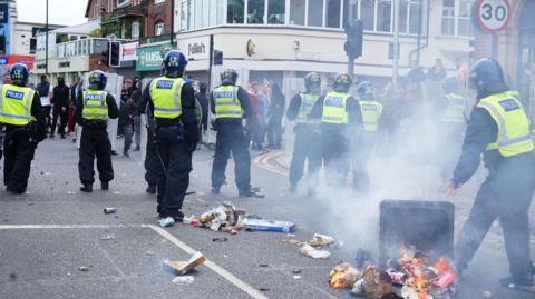 A number of police and protesters with a bin that is tipped over and on fire