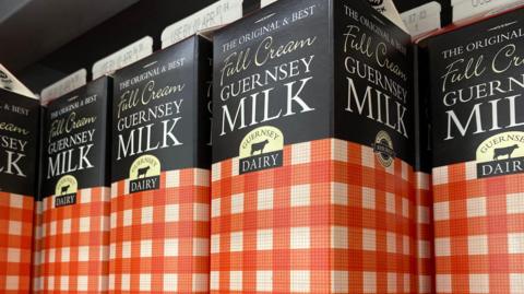 A close-up of four cartons of Guernsey Diary milk. Half of the carton is black, and the other half is orange and white squares.