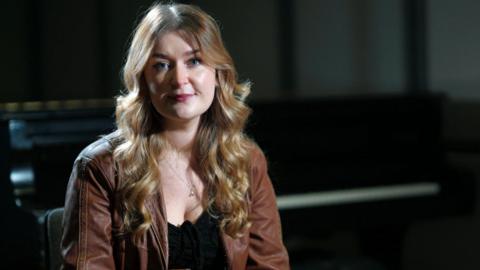 Josie Duncan has long, curly fair hair and is wearing a brown jacket over a black top. She is sitting in a darkened room looking at a camera.