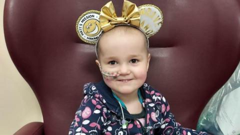 A girl wearing a blue, pink and white hoodie, with a tube in her nose and wearing gold Mickey Mouse ears. She is sat in a large purple armchair.