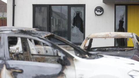 Three burnt-out cars outside a house with a yellow door, which has some broken windows