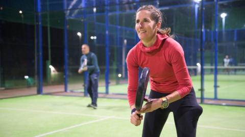 Woman padel tennis player ready to serve on a court at night. She is holding a racquet and leaning forward and smiling. There is a man behind her also ready to play.