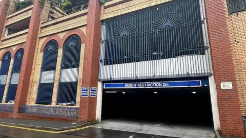 The red and yellow brick Shaw's Brow building with arched window shapes and blue vertical metal bars.