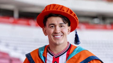 Luke O'Nien is wearing an orange graduation cap (with a dark blue tassel) and is smiling. He is wearing a Sunderland AFC red and white kit underneath his orange graduation gown and seats at the Stadium of Light are out of focus in the background. 