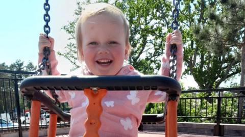 A little toddler on a swing. She has short wispy blonde hair and is beaming at the camera.