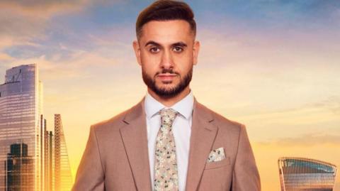 Carlo Brancati is in a beige jacket with a floral tie and matching hankerchief in the pocket. He has a well trimmed beard and black hair. He is standing against a backdrop of the London skyline.