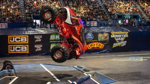 A red monster truck flying through the air as it jumps over a large ramp in an arena, where thousands of people are watching from the stands. The truck is red and designed like Spider-Man, with black spider web patterns and a large spider on the roof.