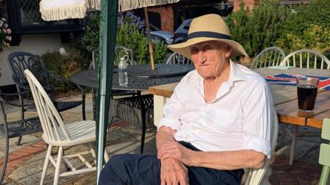 A man in a white shirt and black trousers wearing a sun hat and sitting outside in the sun.