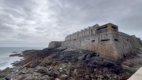 A large stone coastal castle sits on a rocky shoreline beside rough, grey-blue sea. Thick defensive walls with small rectangular openings run along the structure, and a long building with dormer windows stretches across the top. Waves gently lap against the rocks below under an overcast, cloudy sky.