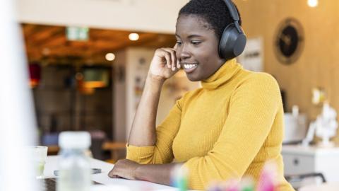 A woman wearing a yellow jumper and black headphones smiling while looking at a computer screen