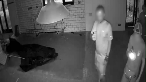 A black and white CCTV image of two men in an abandoned building. One is taking photos on his phone and the other appears to be shining a torch.