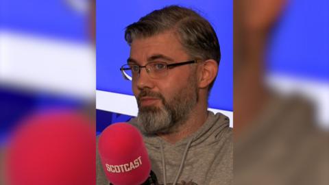 A man with ashy brown hair, a grey beard and blue eyes is wearing black glasses and a grey hoodie. He is sitting in front of a purple background and speaking into a red Scotcast mic.