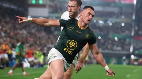 South Africa's Jesse Kriel wheels away and spreads his arms wide like an aeroplane with fingers pointed after scoring a try in the Nations Championship against Wales. Kriel, who is visible from the knees up, wears a green Springbok jersey with yellow trim and white shorts. One bearded Wales defender in a white shirt is partially hidden just behind Kriel